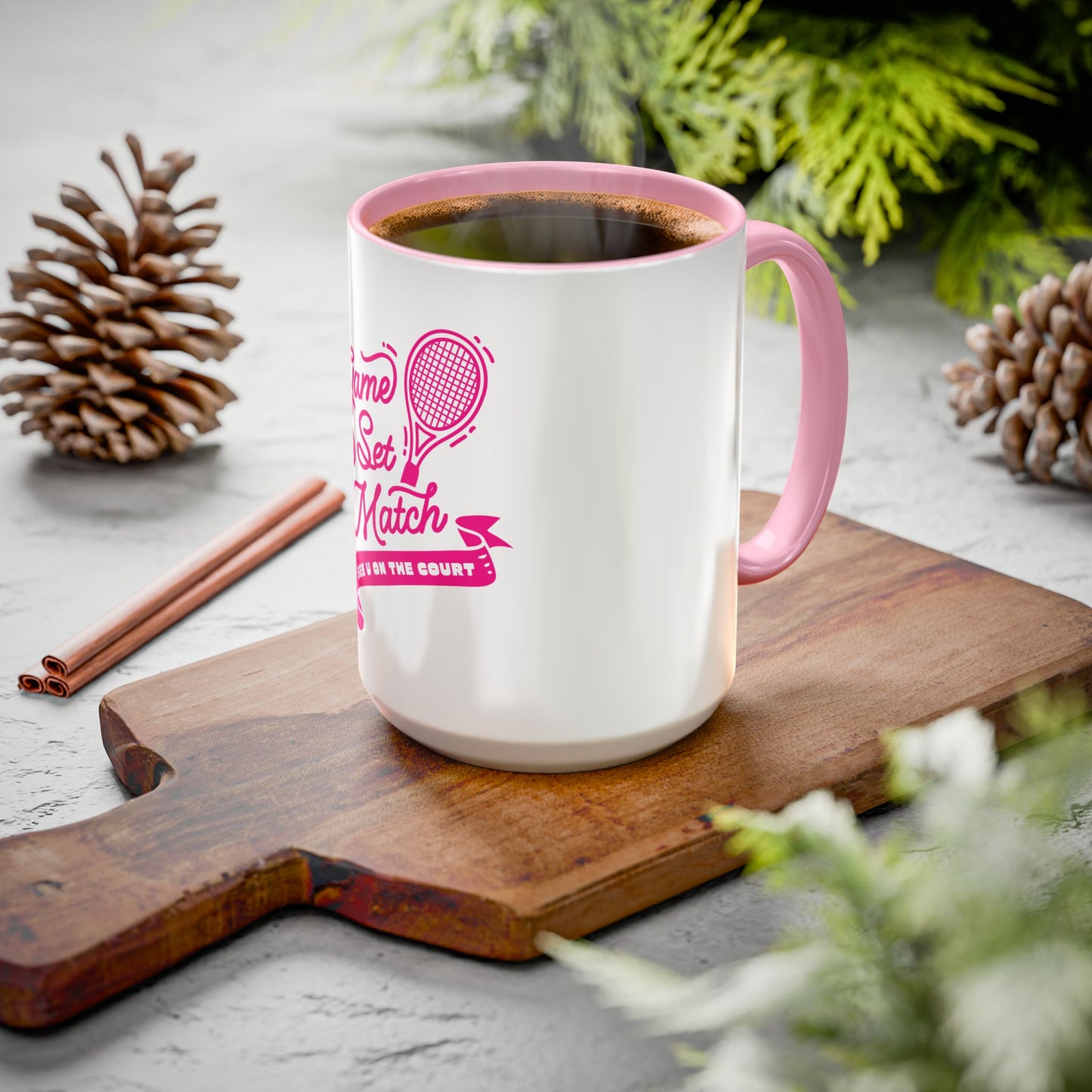 White mug with pink handle and design on a wooden cutting board with pine cones and cinnamon sticks.