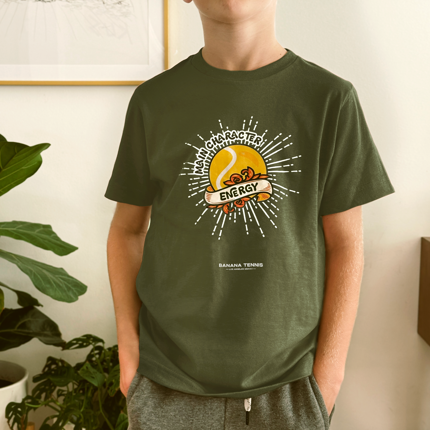 Boy in dark olive green 'Main Character Energy' shirt with yellow tennis ball sunburst design, indoor plant background