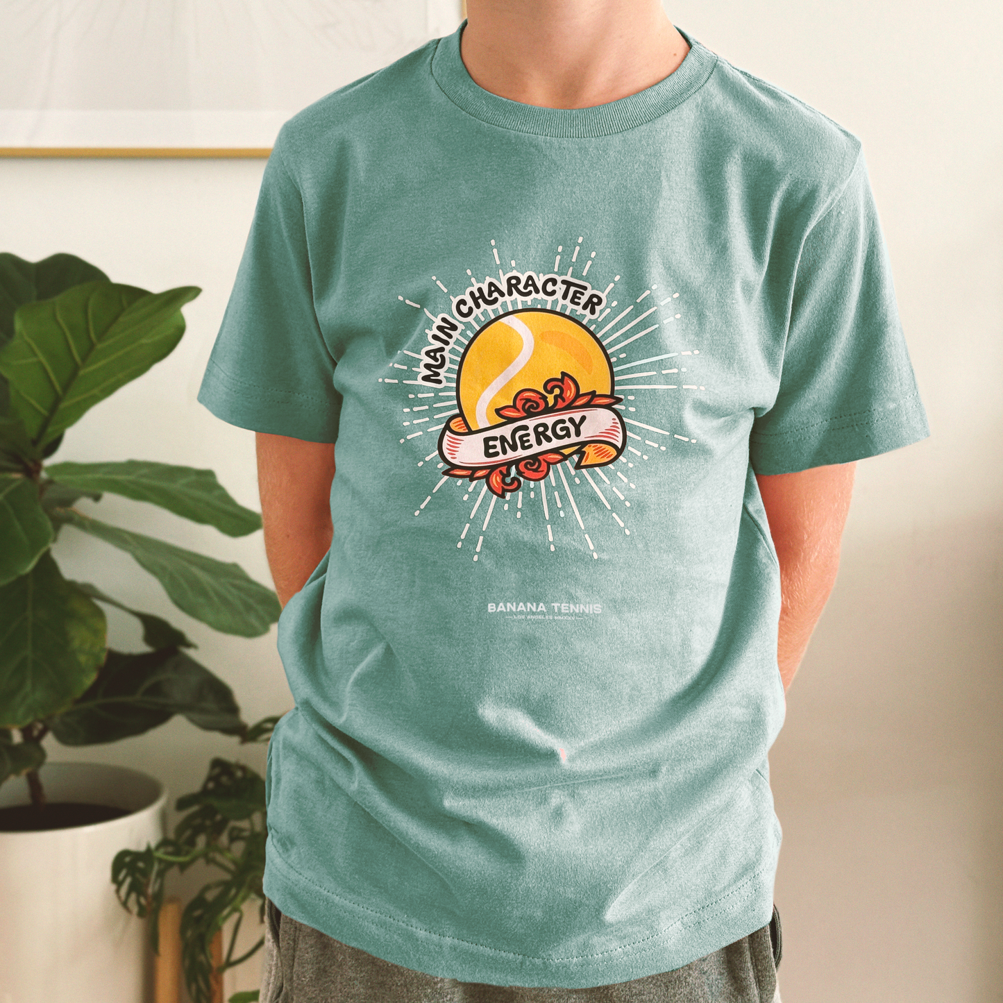 Child in Mint green 'Main Character Energy' tee with tennis-themed sunset design and white burst lines, indoor plant background