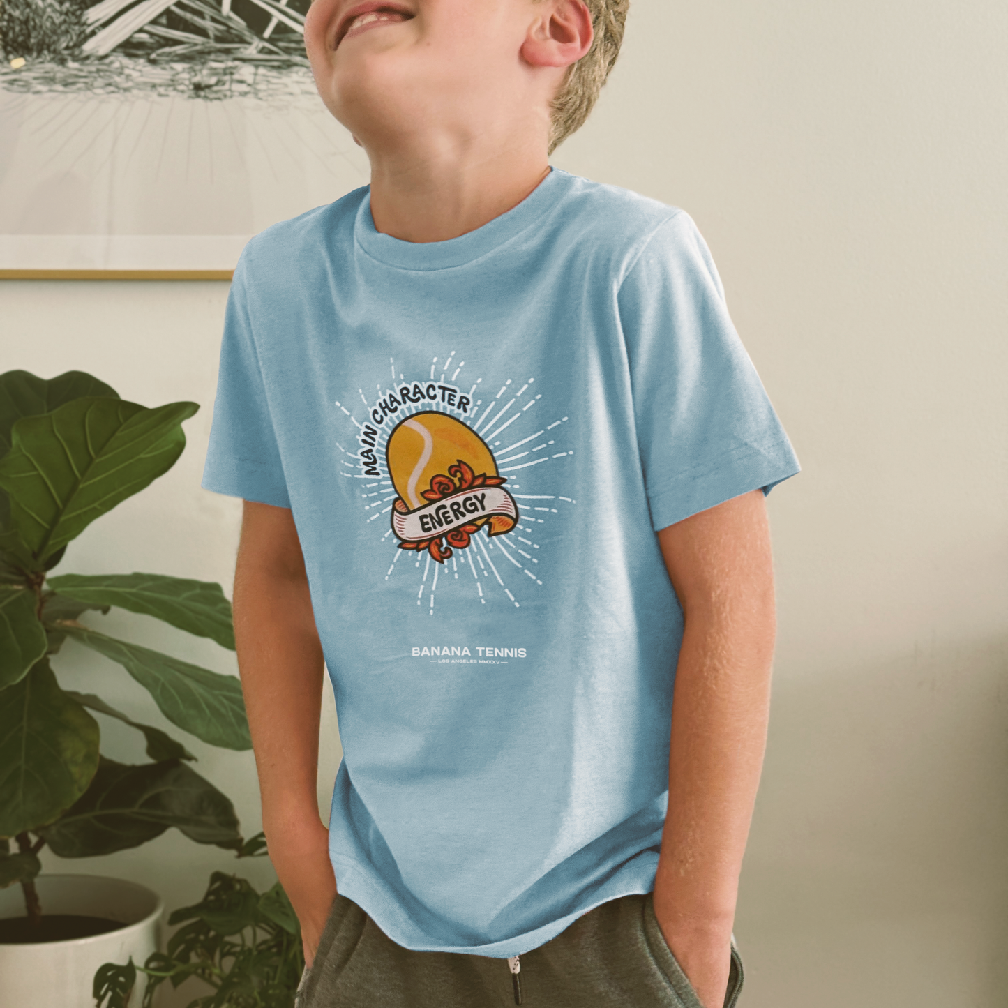 Young boy wearing light blue 'Main Character Energy' tennis t-shirt, smiling with plants in background