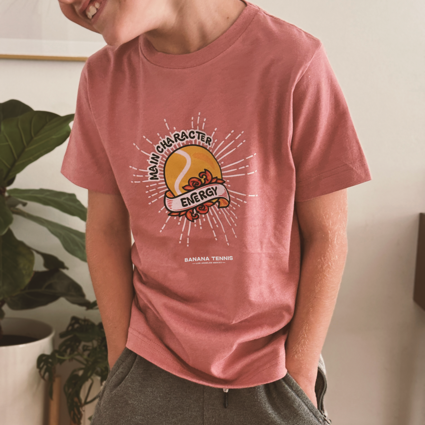 Smiling boy wearing rose pink 'Main Character Energy' t-shirt with retro tennis graphic, standing in bright indoor setting with plants