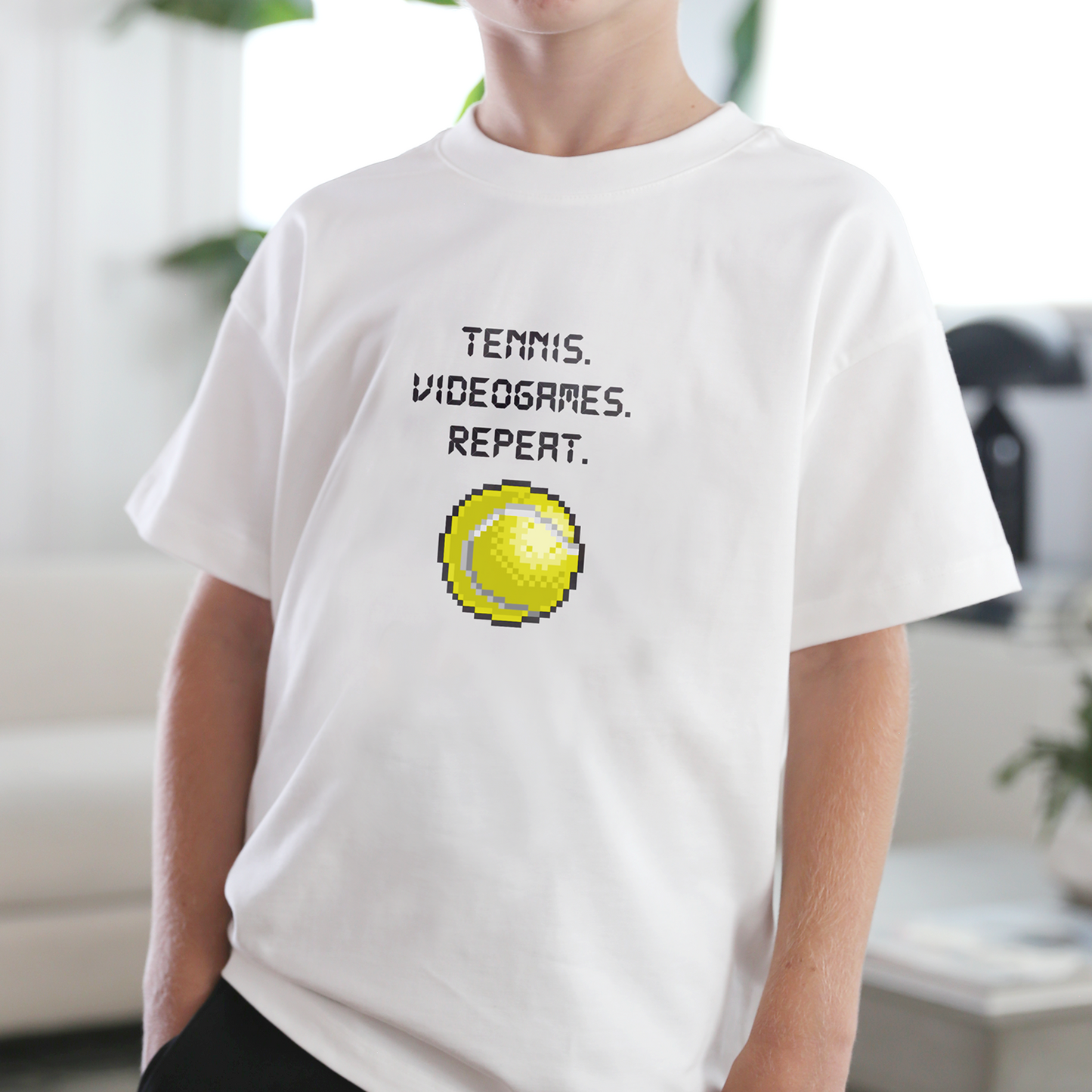 Person wearing a white t-shirt with a bitmap graphic of a pixelated tennis ball and TENNIS. VIDEOGAMES. REPEAT text in bright living room