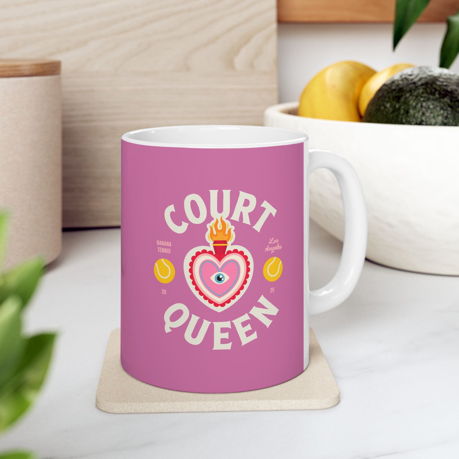 Pink mug with 'Court Queen' design on a white surface with plants and a plant pot in the background