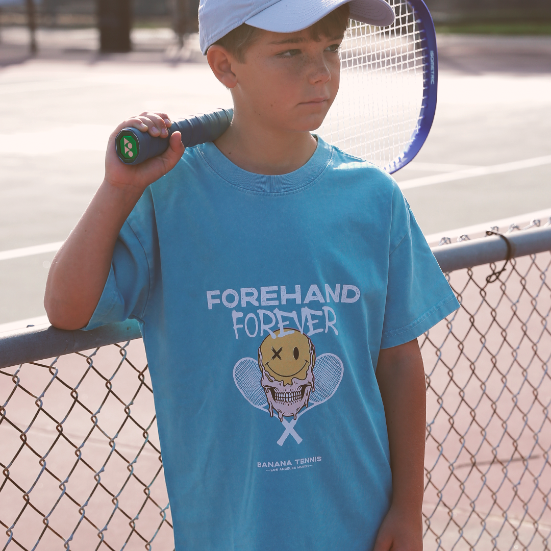 Child wearing a blue t-shirt with a tennis-themed design, holding a tennis racket, standing on a tennis court.