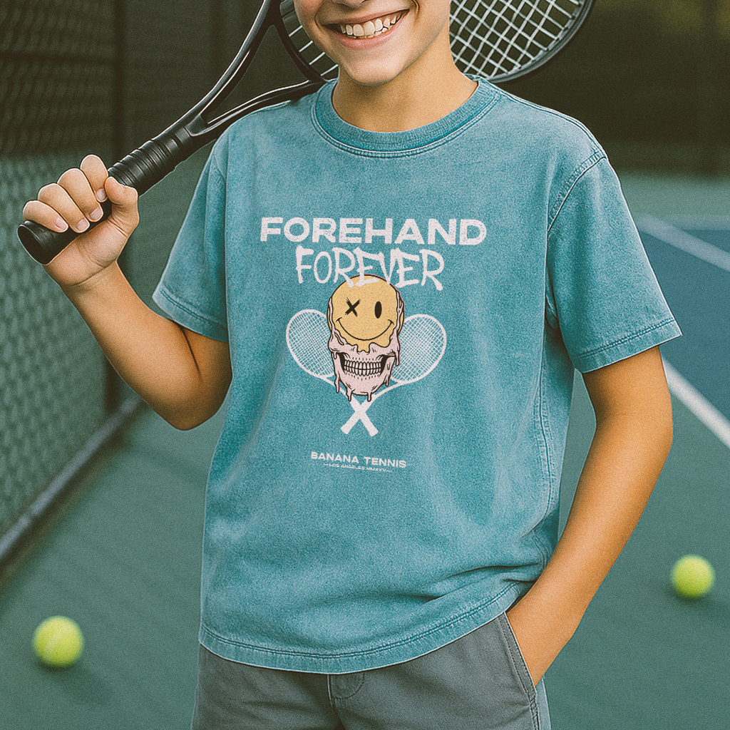 Child wearing a blue t-shirt with a tennis-themed design reading FOREHAND FOREVER on a tennis court
