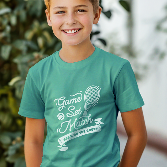 Smiling boy wearing teal 'Game Set Match' youth tennis t-shirt with white text and tennis racket graphic, outdoors with greenery in background