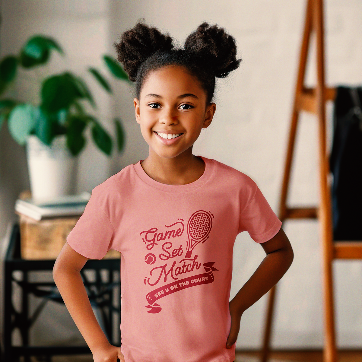 Smiling girl wearing pink youth tennis t-shirt with 'Game Set Match' text and tennis racket graphic, standing indoors