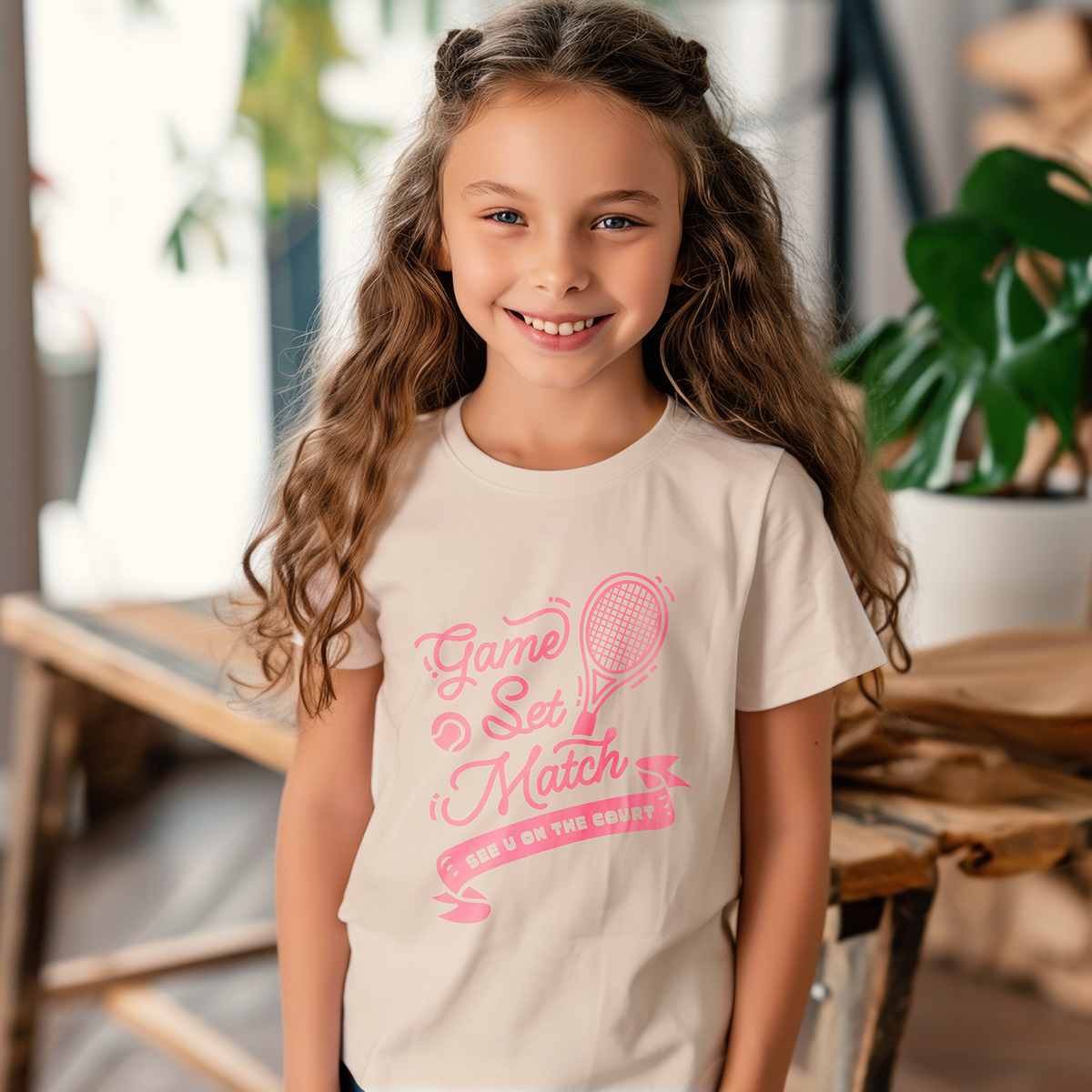 Young girl wearing cream-colored tennis tee with pink 'Game Set Match' print and tennis racket graphic, standing near a window