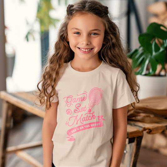 Young girl wearing cream-colored tennis tee with pink 'Game Set Match' print and tennis racket graphic, standing near a window