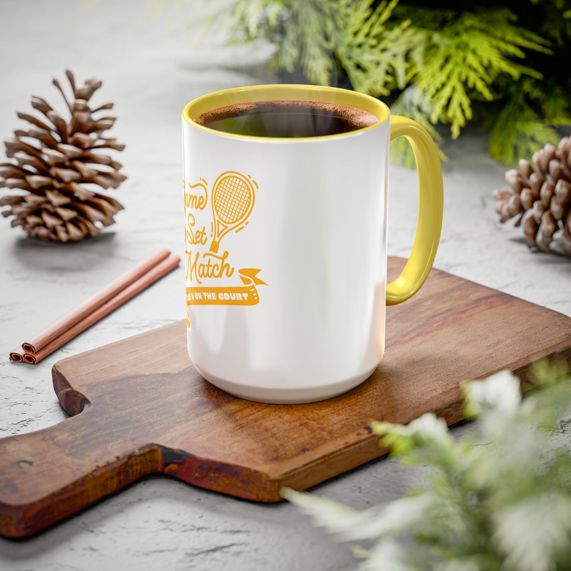 White mug with yellow handle and text on a wooden board with pine cones and cinnamon sticks.