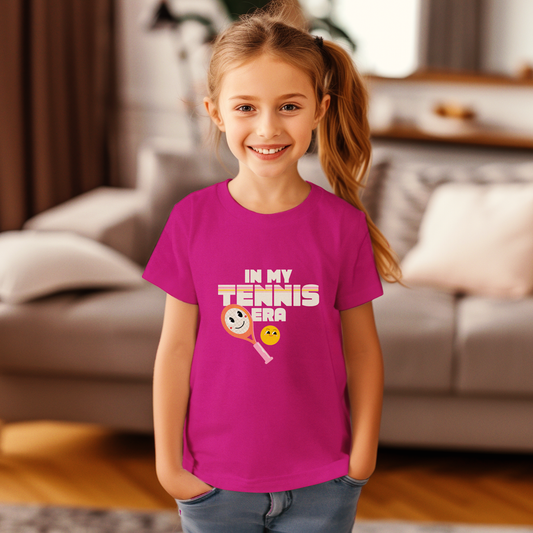 Young girl wearing bold magenta 'IN MY TENNIS ERA' tee with smiling tennis ball icon, posing in cozy indoor setting