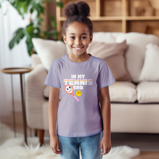Girl wearing lavender 'IN MY TENNIS ERA' t-shirt with tennis emoji graphic, smiling in bright living room