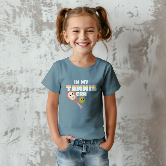 Smiling girl wearing dusty blue 'IN MY TENNIS ERA' t-shirt with graphic tennis ball emoji, standing against textured gray wall
