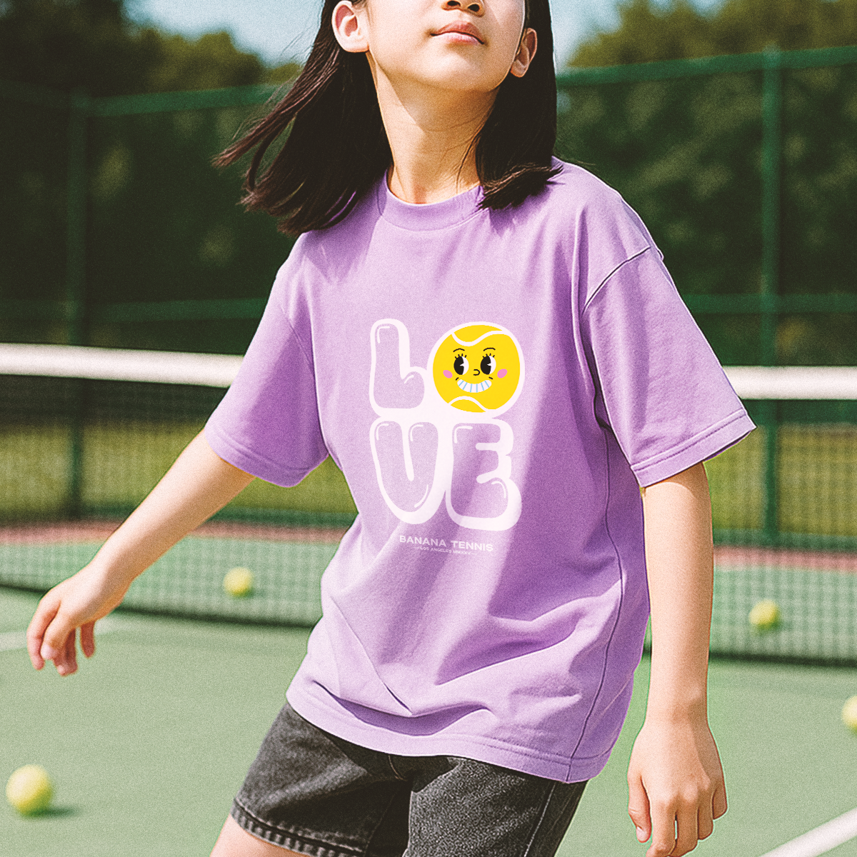 Smiling youth wearing light purple 'LOVE' t-shirt with large tennis ball face design, mid-step on tennis court
