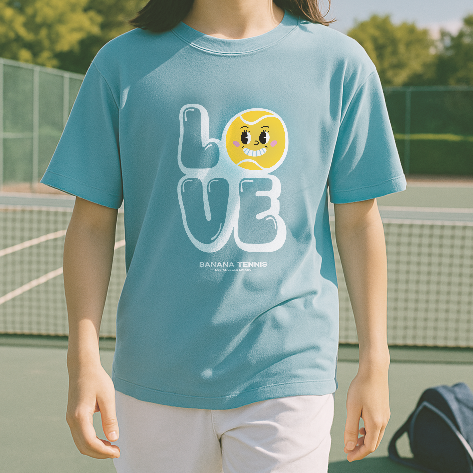 Youth in light blue 'LOVE' tennis t-shirt with bold white lettering and tennis ball face graphic, walking across court