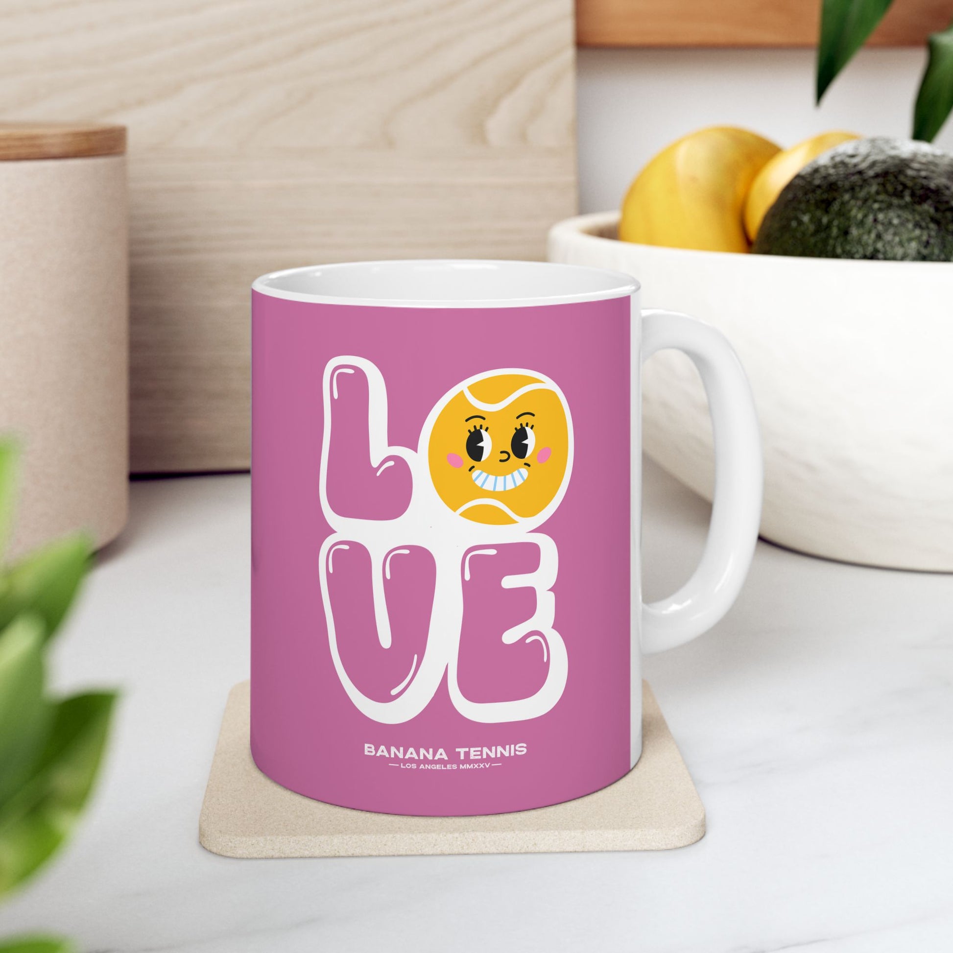 Pink mug with 'LOVE' and banana tennis design on a white surface with plants and a plant pot in the background.