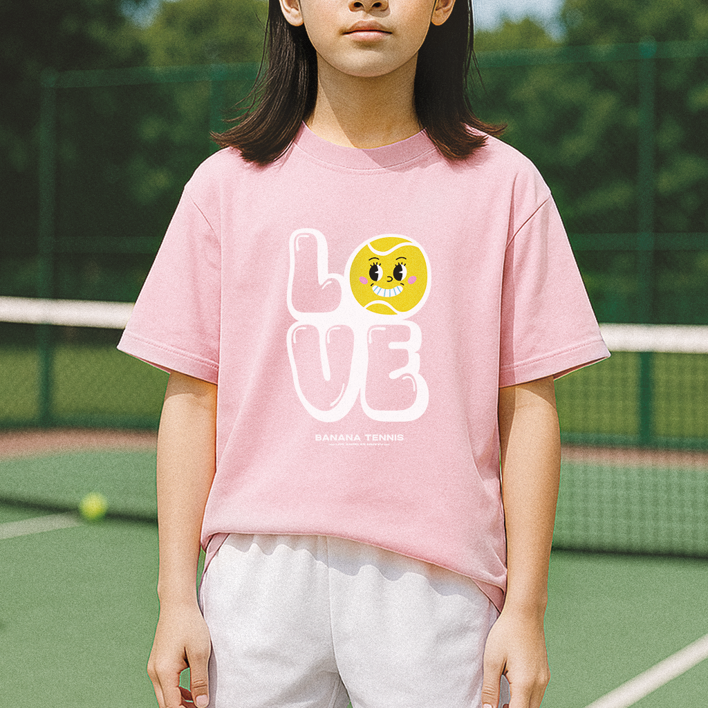 Child standing on court in pastel pink 'LOVE' tennis t-shirt with white letters and cute tennis ball character