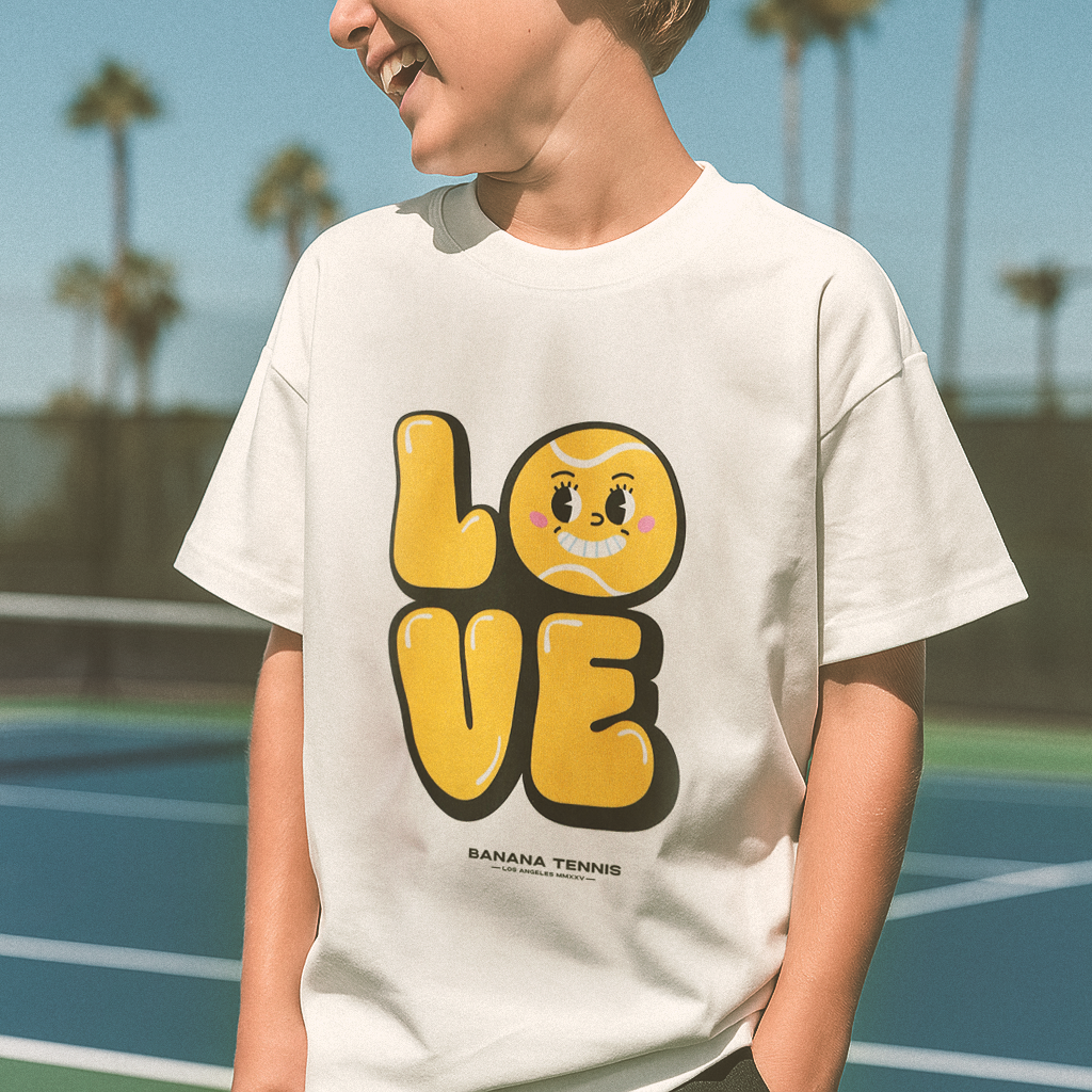 Smiling boy wearing white t-shirt with bold yellow 'LOVE' graphic featuring a smiling tennis ball face as the letter O, standing on sunny outdoor tennis court with palm trees in background
