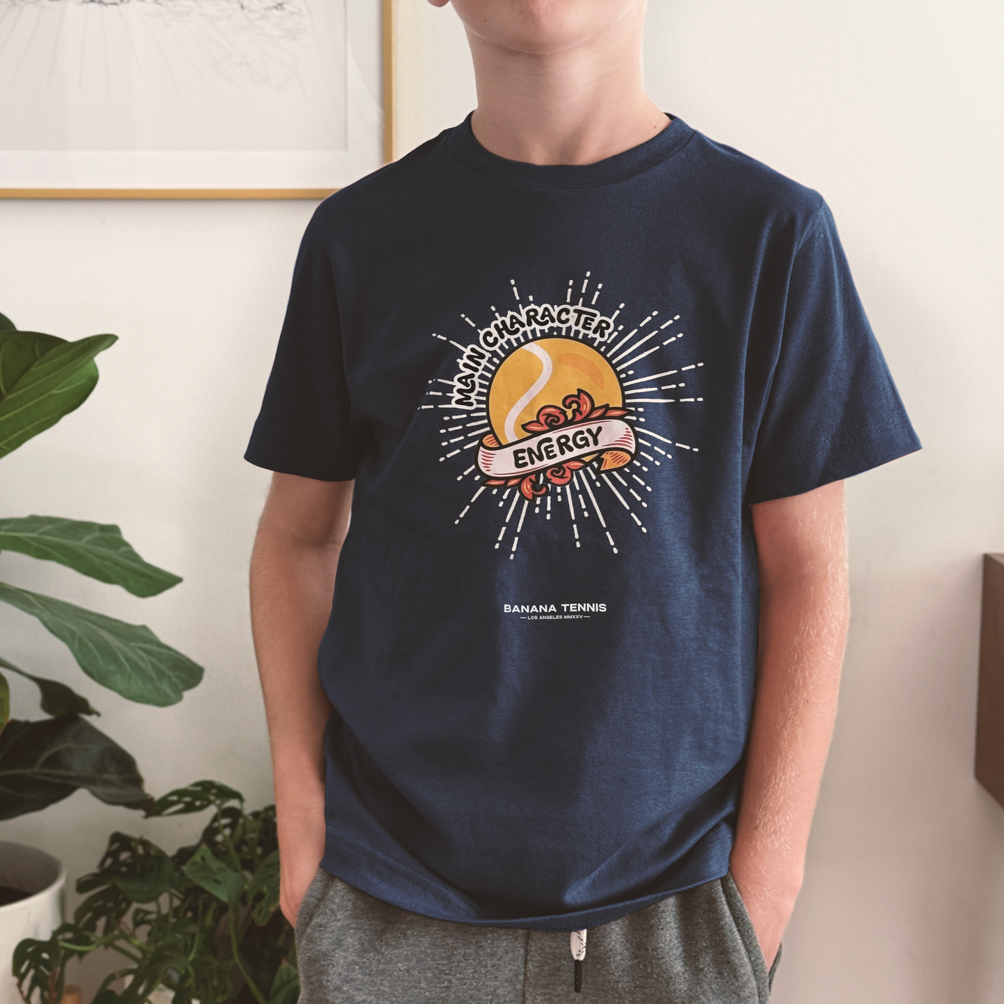Kid wearing navy 'Main Character Energy' youth t-shirt with sunburst tennis ball graphic and bold text, standing indoors
