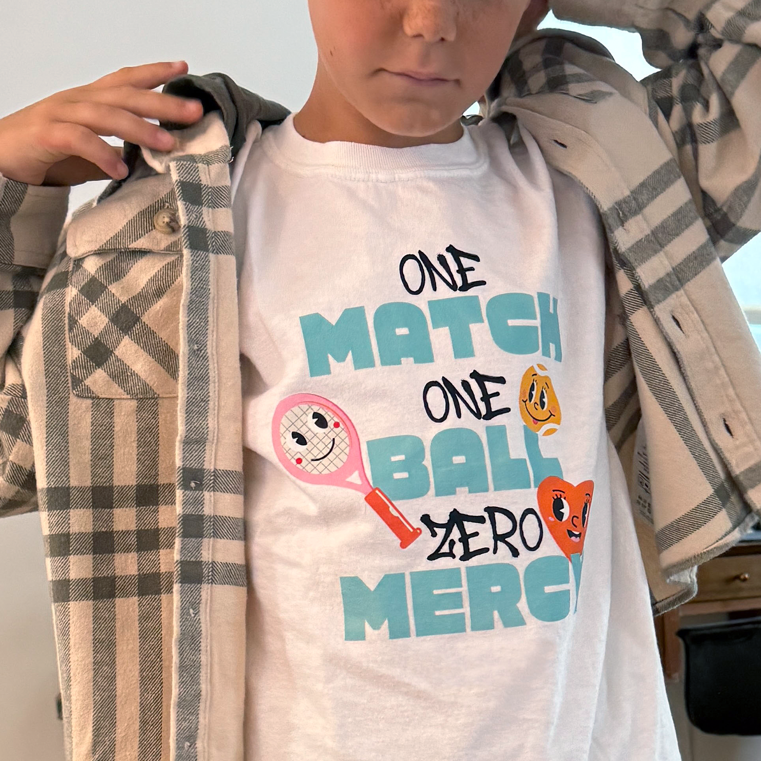 Youth wearing white graphic tennis tee with fun multicolor lettering and cartoon tennis ball illustrations, styled under open plaid shirt