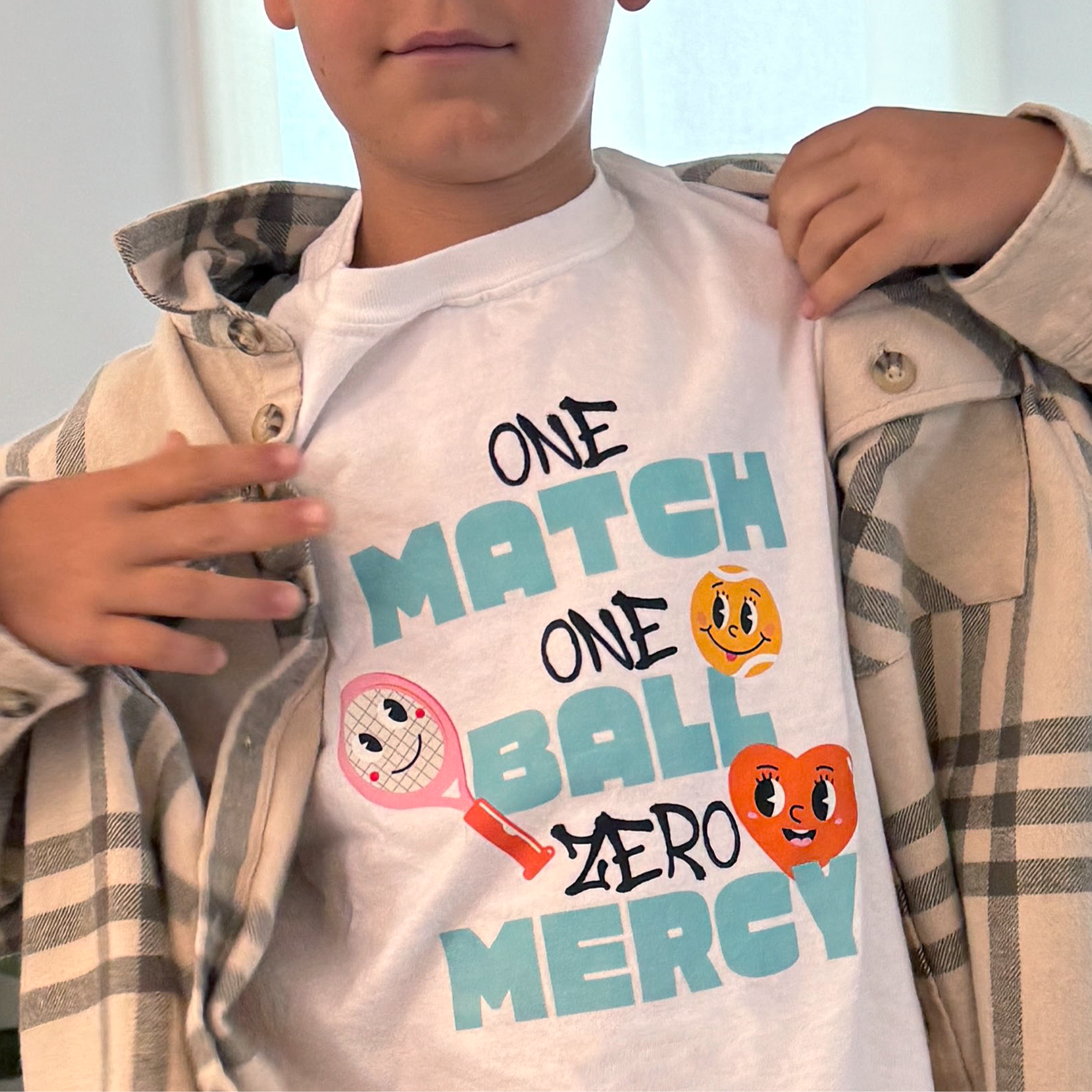Boy opening flannel shirt to reveal white t-shirt with colorful text reading 'ONE MATCH ONE BALL ZERO MERCY' and tennis ball characters