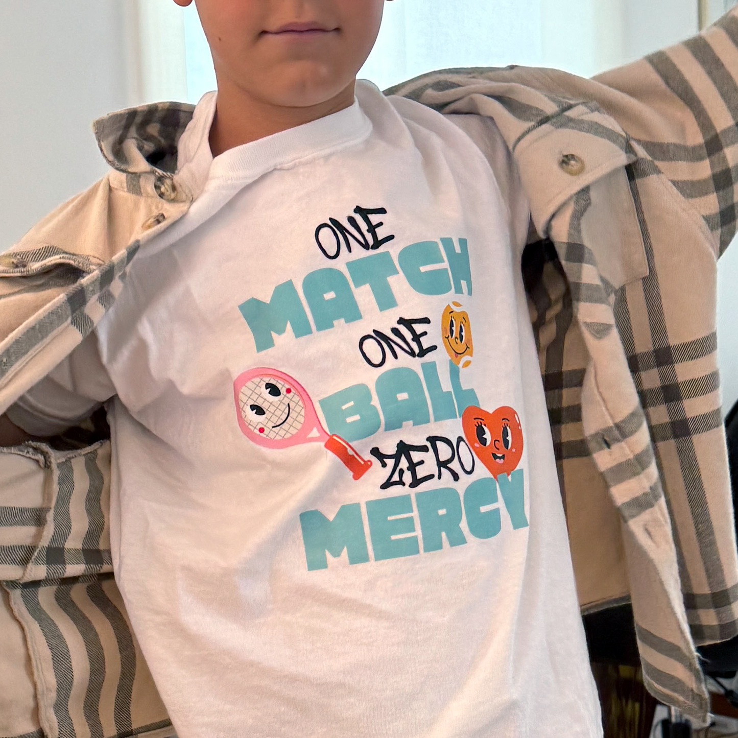 Close-up of white t-shirt with bold 'ONE MATCH ONE BALL ZERO MERCY' design and three expressive tennis ball faces, worn under plaid shirt