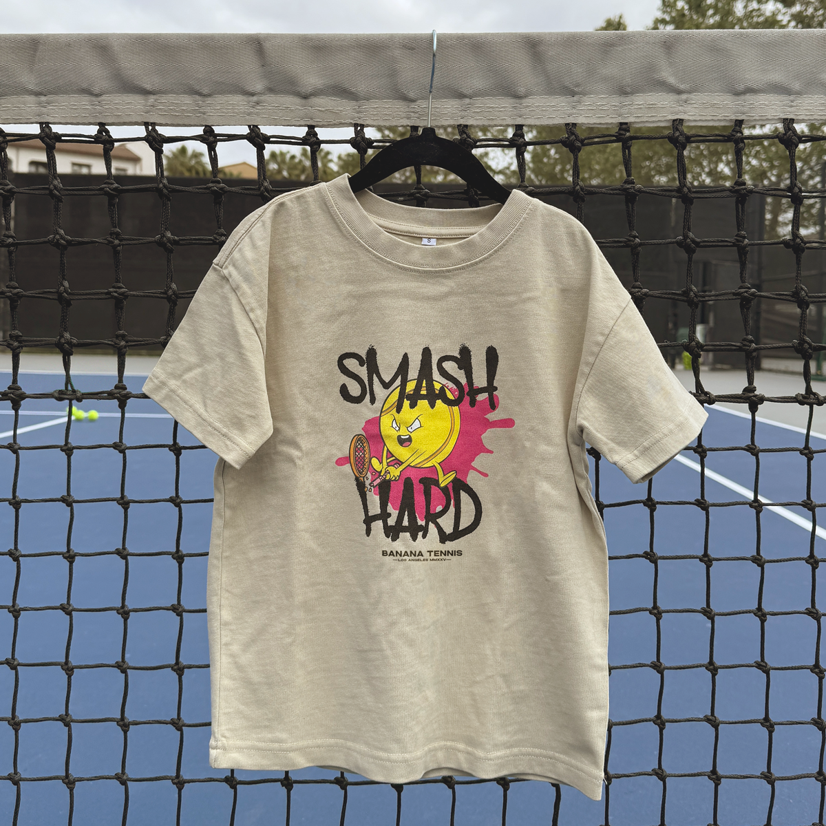 Sand 'SMASH HARD' t-shirt with angry yellow tennis ball graphic hanging on tennis net outdoors