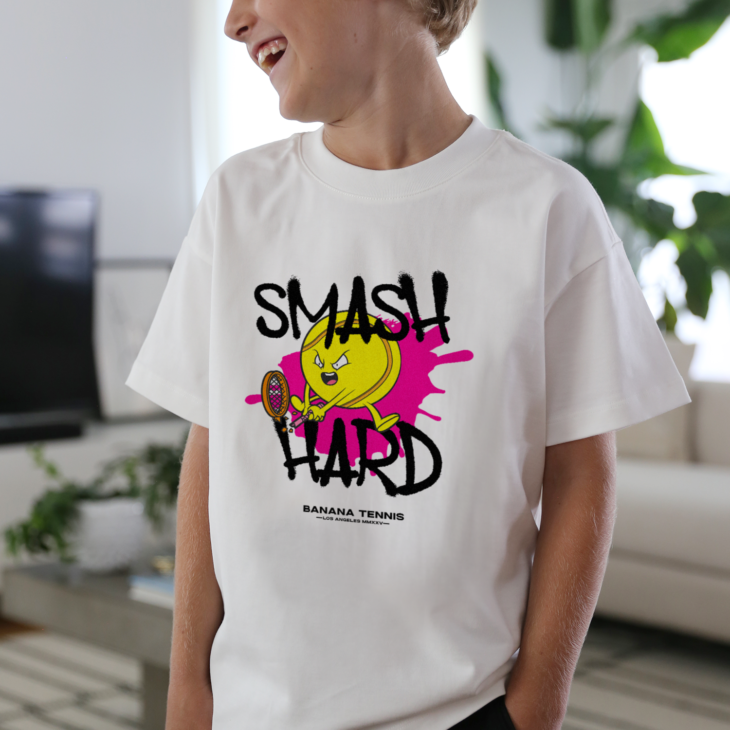 Smiling child wearing white t-shirt with 'SMASH HARD' tennis ball graphic and pink splash, standing in bright living room
