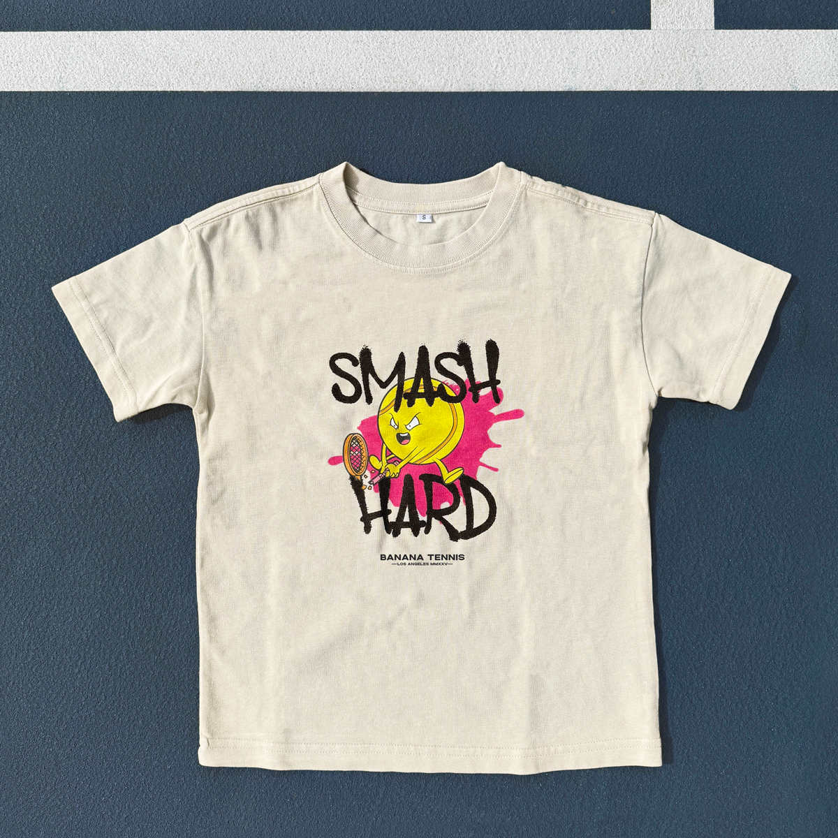 Flat-lay of white 'SMASH HARD' youth t-shirt on blue tennis court surface