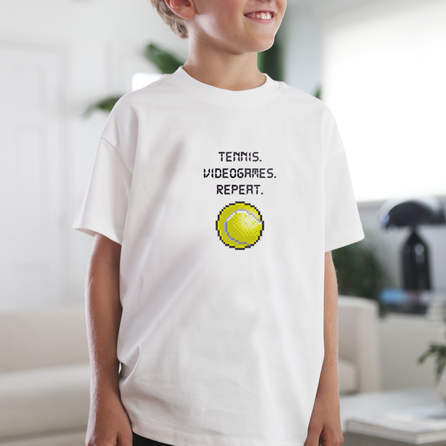 Child wearing a white t-shirt with a bitmap graphic of a pixelated tennis ball and TENNIS. VIDEOGAMES. REPEAT text in bright living room