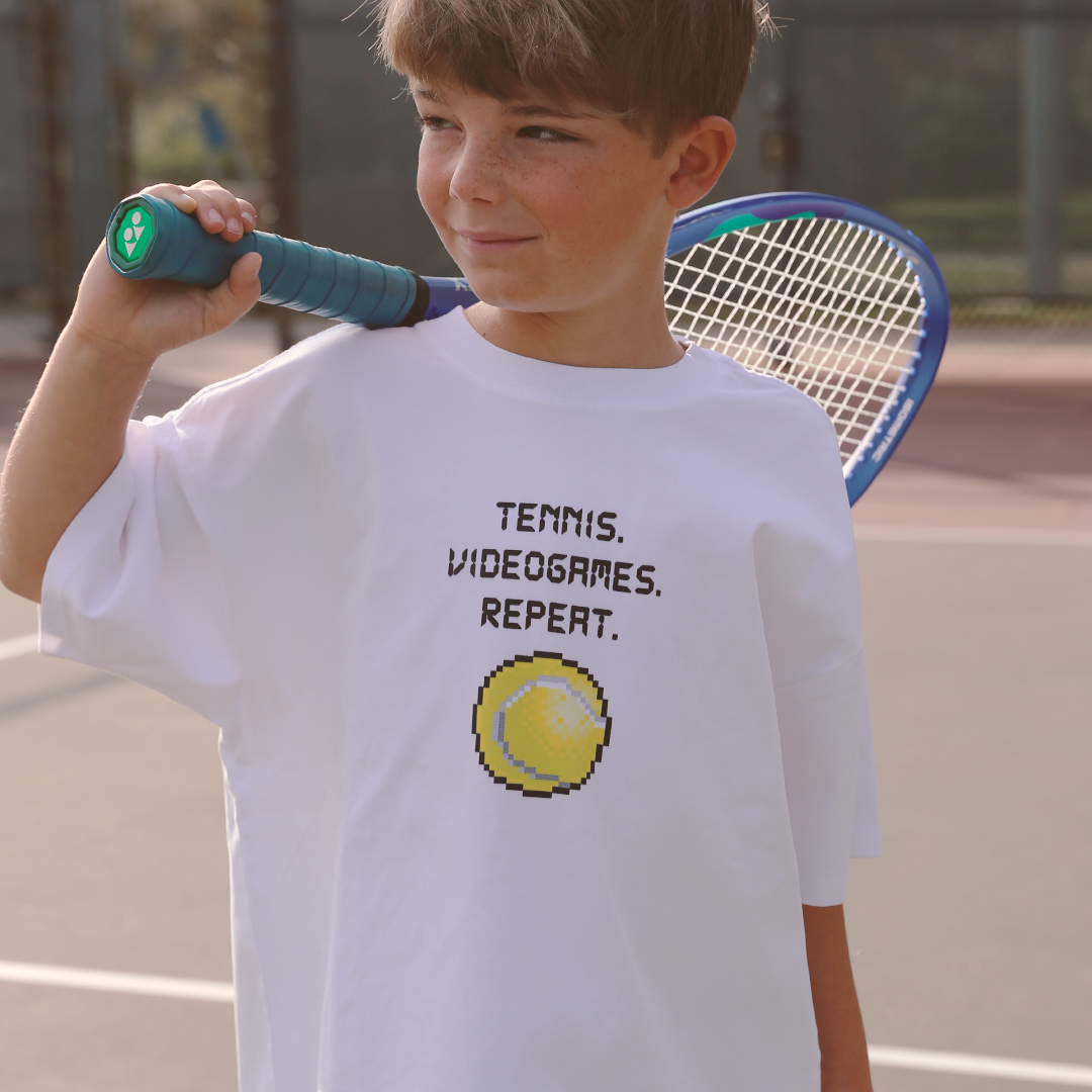 Child wearing a white t-shirt with 'Tennis. Videogames. Repeat.' text and pixelated tennis ball graphic on a tennis court.