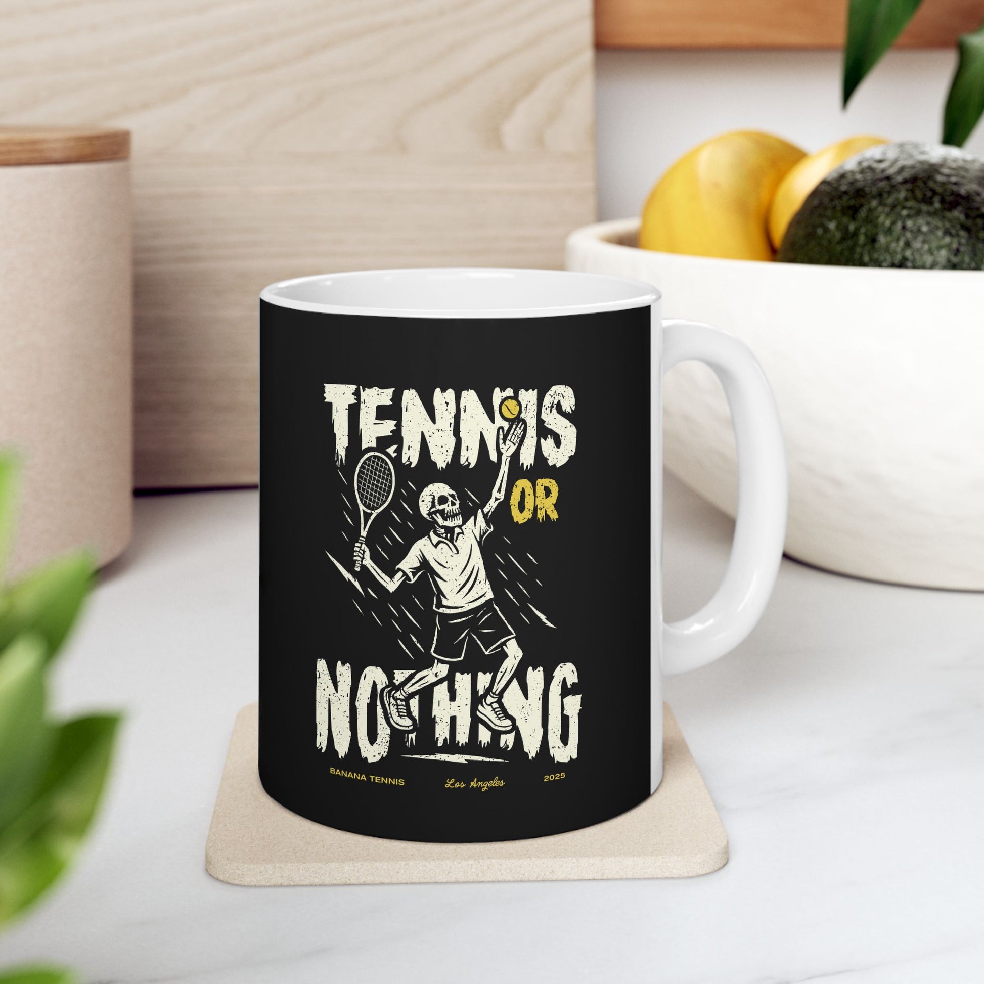 Mug with 'Tennis or Nothing' design on a coaster, placed on a surface with a plant and container in the background.