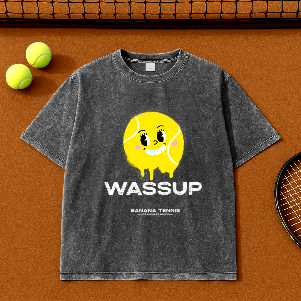 Dark gray WASSUP t-shirt with yellow tennis ball character, flat-lay with tennis balls and racket