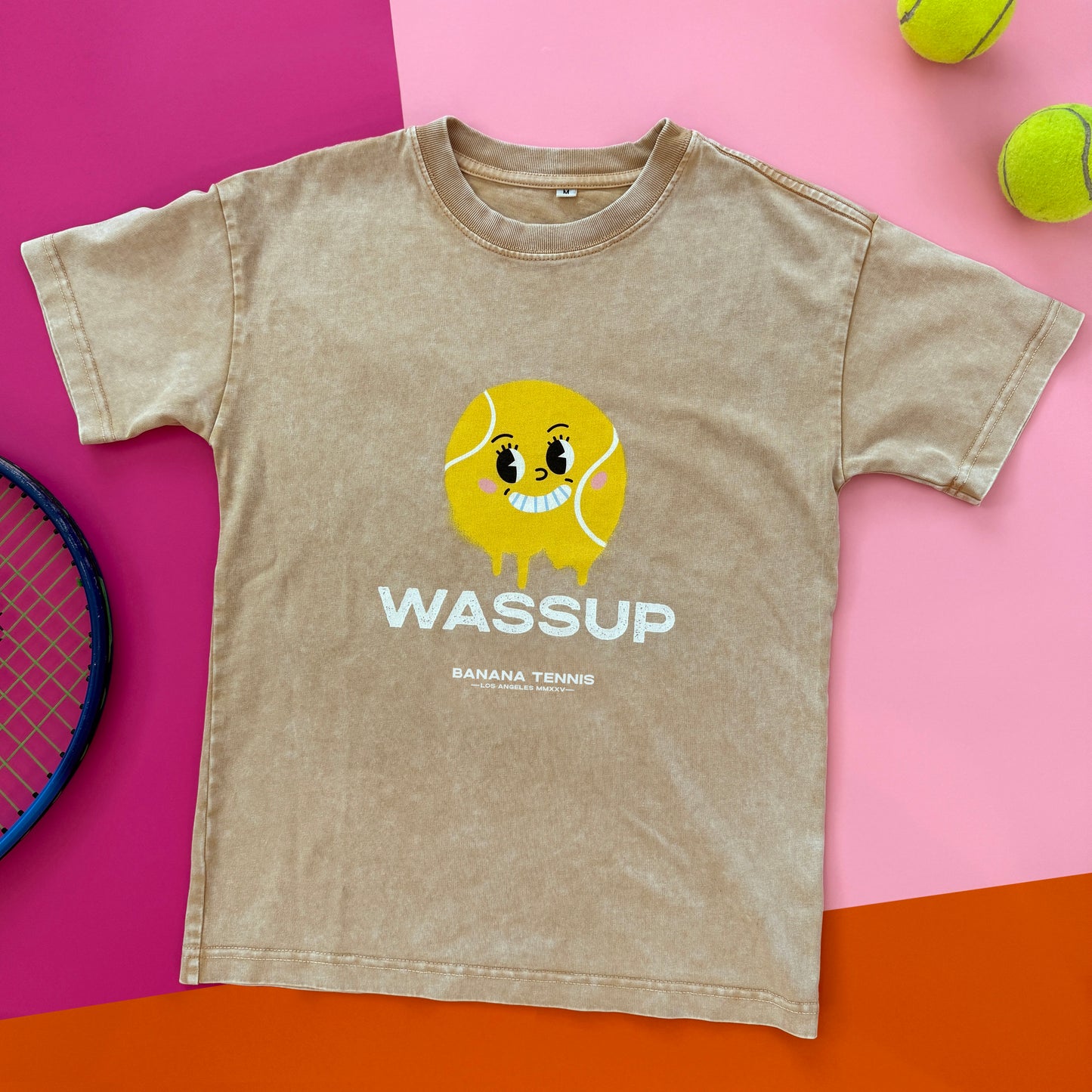 Beige WASSUP youth t-shirt with yellow dripping smiley tennis ball design, flat-lay on bright orange and pink background