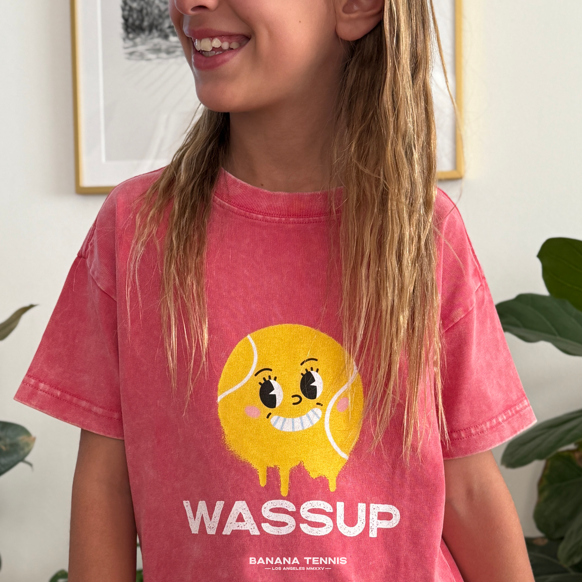 Smiling girl wearing vintage cherry WASSUP tee with big yellow tennis ball face graphic, standing in casual indoor setting