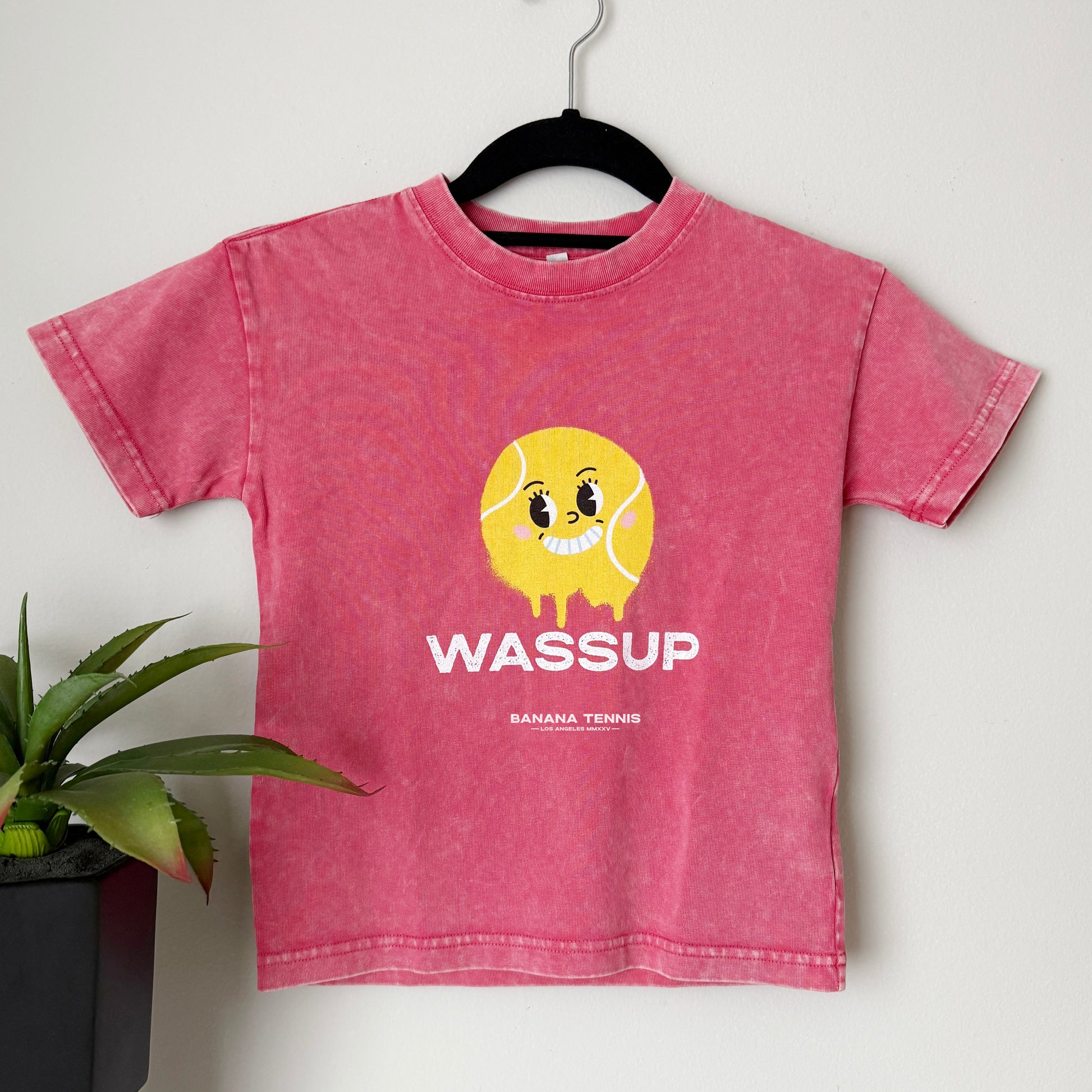 Faded pink WASSUP t-shirt with large yellow tennis ball face graphic, hanging on hanger near plant