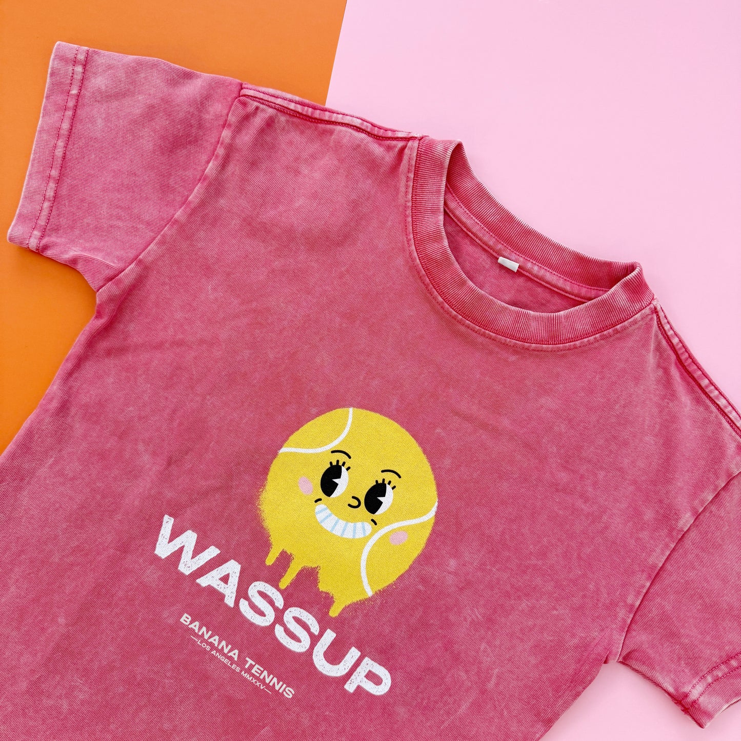 Pink t-shirt with a cartoon tennis ball and 'WASSUP' text on a pink and orange background