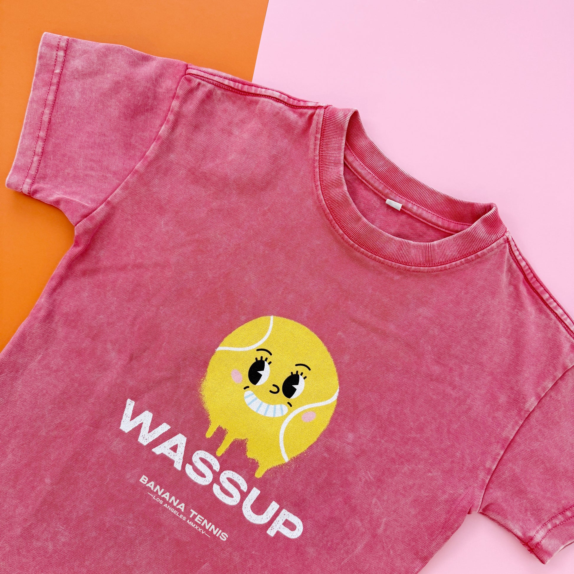 Pink t-shirt with a cartoon tennis ball and 'WASSUP' text on a pink and orange background