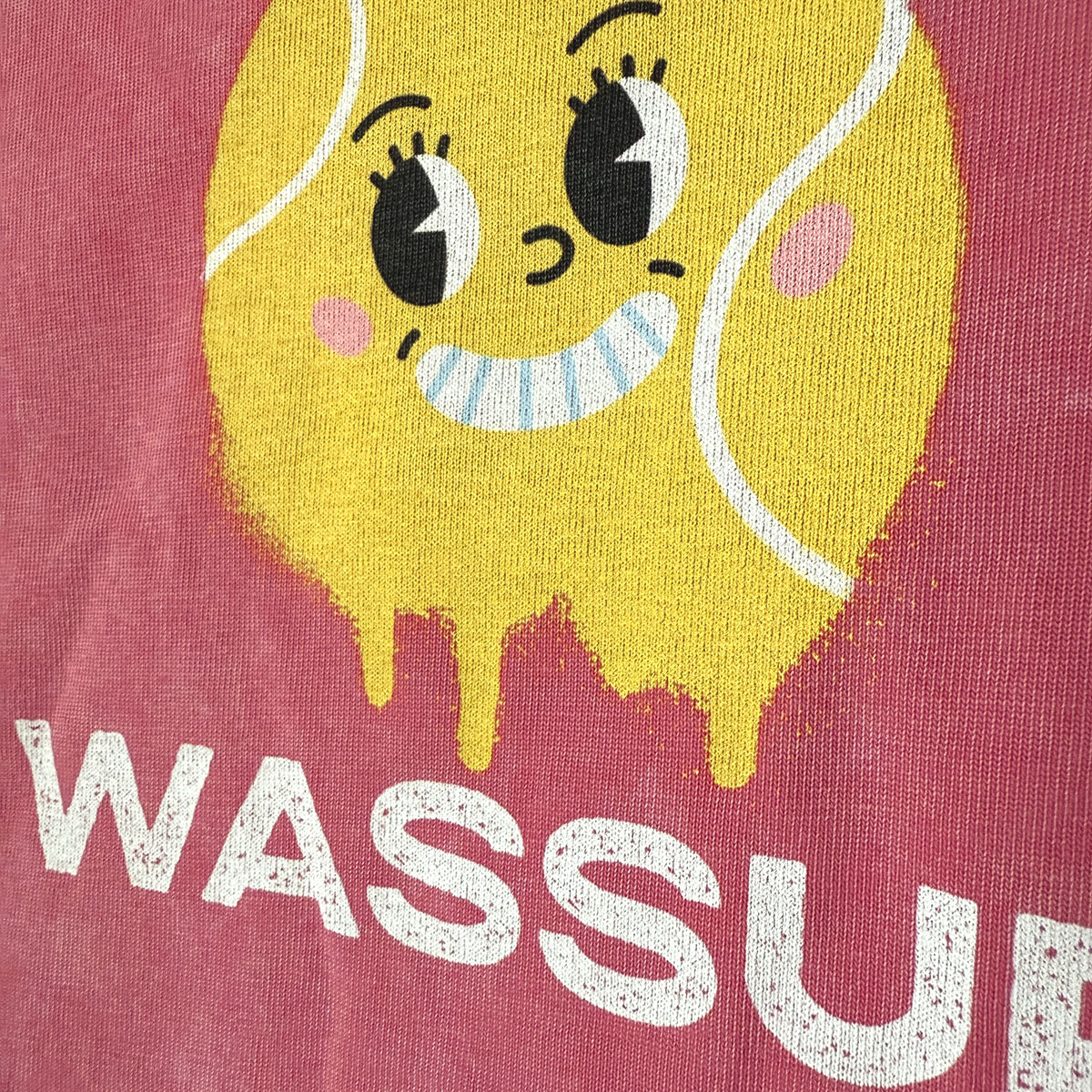 Close-up of red WASSUP t-shirt showing textured yellow smiley face tennis ball graphic with white text
