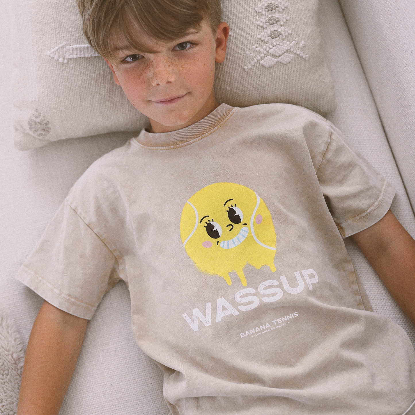 Young boy lying on sofa wearing beige WASSUP t-shirt with large yellow smiley tennis ball graphic, relaxed indoor scene