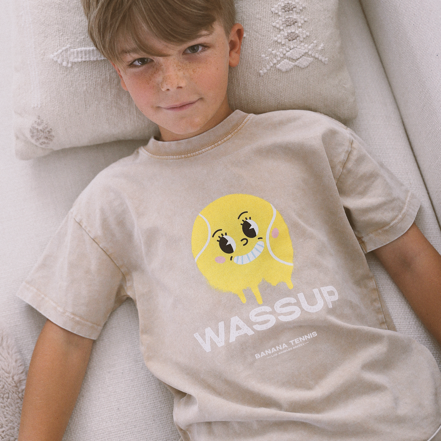 Young boy lying on sofa wearing beige WASSUP t-shirt with large yellow smiley tennis ball graphic, relaxed indoor scene