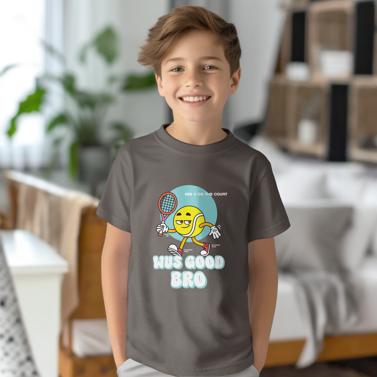 Smiling boy wearing charcoal gray 'WUS GOOD BRO' t-shirt with cartoon tennis ball character and racquet, standing in modern living room